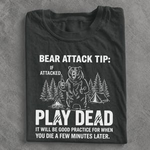 May include: A black crew neck t-shirt featuring a white graphic of a bear holding a bottle near a campfire and tent. The text reads: BEAR ATTACK TIP: IF ATTACKED, PLAY DEAD. IT WILL BE GOOD PRACTICE FOR WHEN YOU DIE A FEW MINUTES LATER.