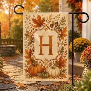 May include: A rectangular garden flag featuring a cream-colored background with a central letter H in a serif font. The design is framed by a border of autumn leaves, acorns, and three pumpkins in orange, white, and green at the bottom.
