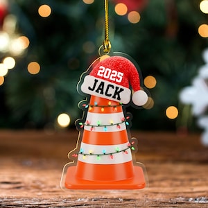 May include: A flat, hanging ornament shaped like an orange and white traffic cone wrapped in colorful string lights. A red Santa hat sits on top of the cone, featuring the white text 2025 and the name JACK in bold black capital letters.