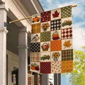 May include: A rectangular garden flag featuring a patchwork design with autumn-themed squares. Patterns include pumpkins, sunflowers, a bird, a green truck, a basket of flowers, a watering can, plaid, polka dots, and maple leaves in orange, red, and green.
