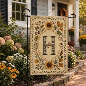 May include: A rectangular garden flag featuring a large, dark green letter H centered within an ornate border. The design includes illustrations of sunflowers, autumn leaves, and acorns in shades of yellow, orange, brown, and green on a beige background.