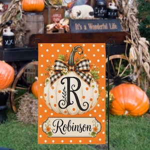May include: A rectangular garden flag with an orange background featuring white polka dots. A large white pumpkin sits in the center, adorned with a black and white buffalo plaid bow and the letter R. Below, a label displays the name Robinson.