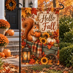 May include: A rectangular garden flag featuring a brown squirrel holding a small pumpkin, sitting on a wooden cart filled with sunflowers, pumpkins, and a plaid blanket. A lit lantern sits nearby, and the text Hello Fall is printed on a wooden sign.
