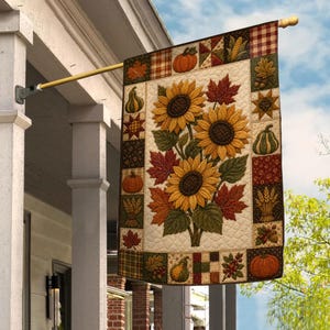 May include: A rectangular decorative flag hanging from a gold metal pole, featuring a central illustration of three yellow sunflowers and red maple leaves on a cream background, surrounded by a border of squares with pumpkins and autumn motifs.