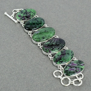 Pode incluir: Uma pulseira de elos de prata com seis pedras de zoisite ovais com inclusões verde-escuras e pretas profundas, acentuadas por pequenas manchas magenta. A pulseira é fixada com um fecho de barra de prata e uma série de anéis circulares.
