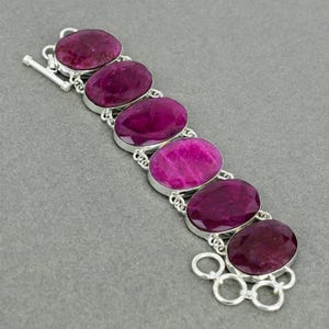 May include: A bracelet featuring six large, oval-shaped, deep magenta-coloured faceted gemstones set in silver-toned metal bezels. The stones are connected by silver-toned metal links, and the piece includes a toggle clasp closure at one end.