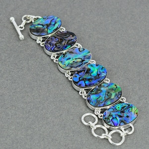 May include: A silver link bracelet featuring six oval-shaped abalone shell cabochons. Each shell displays iridescent swirls of blue, green, and purple. The bracelet is secured with a silver toggle clasp and includes a series of circular chain links.