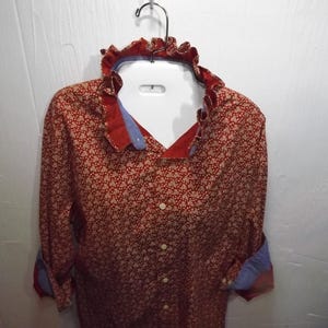 May include: A long-sleeved button-down shirt in a deep red color with a small white floral pattern. The shirt features a ruffled collar, a front button placket, and contrasting light blue fabric visible on the inner collar and sleeve cuffs.
