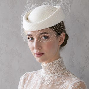 Peut inclure: Un chapeau pillbox blanc doté d'une calotte arrondie structurée et d'un bord plat. Le chapeau est orné d'un voile de type voilette en maille fine et délicate qui s'étend sur le devant, créant une texture douce et superposée sur le visage.