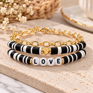 Puede incluir: Un conjunto de pulseras superpuestas que incluye una cadena dorada y dos hileras de cuentas de disco blancas y negras. Una hilera tiene un dije de corazón dorado, mientras que la otra presenta cuentas blancas con letras negras L-O-V-E.