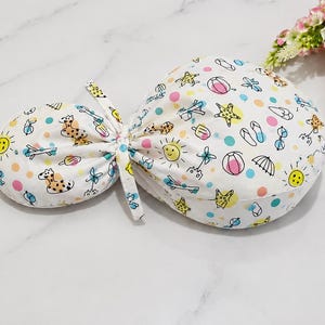 Può includere: Una cuffia per capelli in tessuto bianco con una stampa estiva colorata raffigurante giraffe, soli, stelle marine, infradito, gelati e palloni da spiaggia, arricchita da pois rosa e turchesi sparsi e una chiusura a laccio centrale.