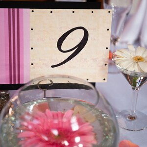 May include: A table setting with a decorative number nine table card. The card has a pink striped design on the left and a beige background with the number nine in black. A white daisy floats in a martini glass.