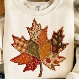 May include: A cream-colored crewneck sweatshirt featuring a large, central maple leaf graphic. The leaf is composed of various fabric patches in autumn colors including orange, brown, green, and tan, with visible decorative stitching along edges.