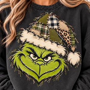 May include: A charcoal gray crewneck sweatshirt featuring a large, central graphic of a green, frowning character wearing a patchwork Santa hat. The hat is composed of green plaid, black and white buffalo check, and leopard print fabric patches.