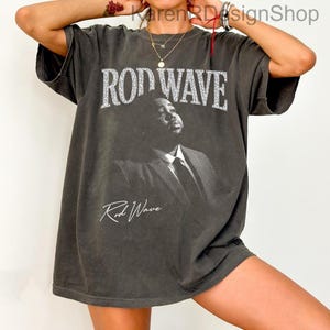 May include: A charcoal gray short-sleeve t-shirt featuring a large black and white photographic print of a person in a suit. The text ROD WAVE is printed in a bold, distressed white font above the image, with the signature Rod Wave below.