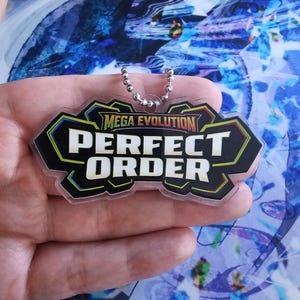 May include: A clear acrylic keyring featuring a black, angular, multi-layered emblem with a green border. The text "MEGA EVOLUTION" appears in small yellow letters above the bold, white, capitalised words "PERFECT ORDER" with a silver ball chain.