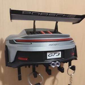 May include: A silver and black wall-mounted key holder shaped like the rear of a Porsche GT3 sports car. The model features a prominent rear spoiler with the text PORSCHE, a red taillight strip, dual exhaust pipes, and a GT3 license plate.