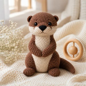 Puede incluir: Un peluche de nutria tejido a ganchillo con cabeza, espalda y extremidades de color marrón oscuro y un pecho y hocico en color crema. El juguete tiene pequeños ojos negros de seguridad, una nariz negra bordada y bigotes blancos, sentado.