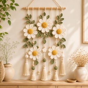 Könnte beinhalten: Ein Wandbehang mit fünf gehäkelten weißen Gänseblümchen mit gelben Mitten und grünen Blättern, die an einer horizontalen Holzstange an cremefarbenen Schnüren hängen. Das Design umfasst Holzperlen und drei cremefarbene Quasten aus Garn.
