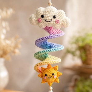 Könnte beinhalten: Eine gehäkelte Hängedekoration mit einer cremefarbenen, lächelnden Wolke oben und einer gelben, lächelnden Sonne unten. Verbunden werden beide durch einen spiralförmigen, regenbogenfarbenen Schweif, der von Rosa zu Violett, Blau, Grün und Gelb verläuft.