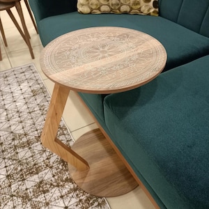 May include: A round wooden side table with a light brown finish and a white engraved mandala pattern on the tabletop. The table features a single angled support leg connected to a circular base, positioned next to a dark green velvet sofa.