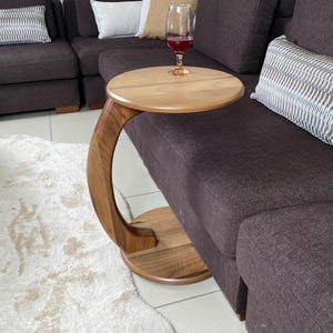 May include: A C-shaped side table crafted from light brown wood with a circular top and a matching circular base connected by a curved support arm. The table features a smooth, polished finish and is positioned next to a dark brown sofa.