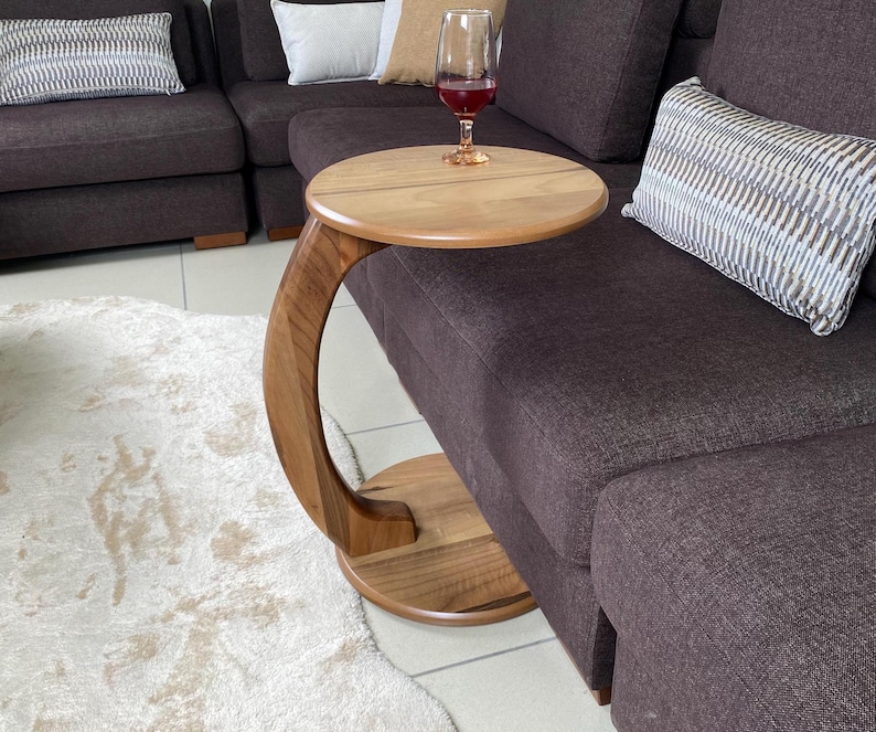 May include: A C-shaped side table crafted from light brown wood with a circular top and a matching circular base connected by a curved support arm. The table features a smooth, polished finish and is positioned next to a dark brown sofa.