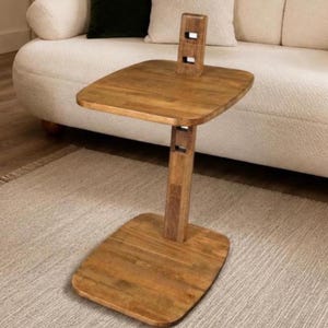 Può includere: Un tavolino a forma di C realizzato in legno di tonalità media con venature visibili. Il tavolo presenta una base rettangolare piatta, un montante verticale con due aperture quadrate e un piano rettangolare coordinato con angoli arrotondati.