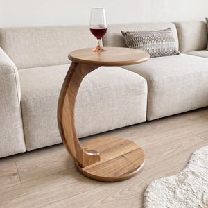 May include: A C-shaped side table crafted from light brown wood with a circular top and a matching circular base. The table features a curved support column connecting the top and base. A clear glass filled with red wine sits on the table surface.