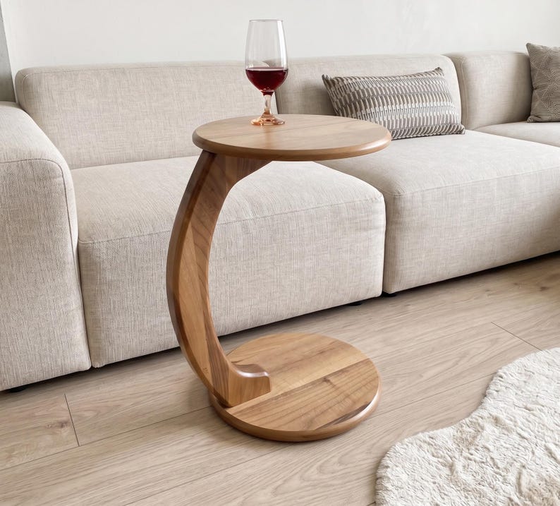 May include: A C-shaped side table crafted from light brown wood with a circular top and a matching circular base. The table features a curved support column connecting the top and base. A clear glass filled with red wine sits on the table surface.