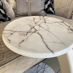 May include: A round side table featuring a white marble top with prominent black veining. The table has a smooth, glossy finish and a curved, solid white base that supports the circular tabletop, creating a clean and functional furniture piece.