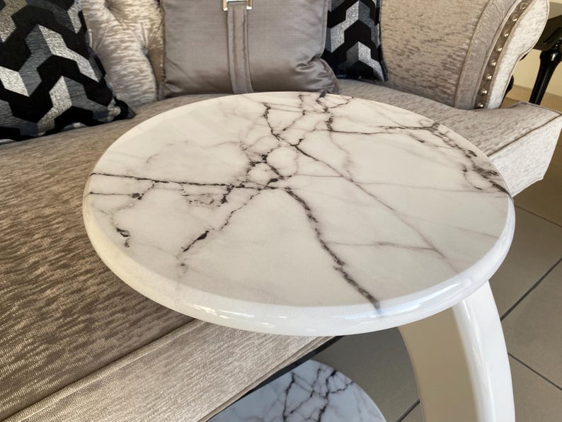 May include: A round side table featuring a white marble top with prominent black veining. The table has a smooth, glossy finish and a curved, solid white base that supports the circular tabletop, creating a clean and functional furniture piece.