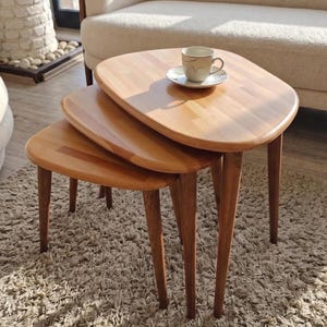 May include: A set of three light brown wooden nesting tables with rounded, irregular shapes and tapered legs. The largest table holds a white ceramic coffee cup on a matching saucer. The tables are arranged in a cascading, overlapping formation.