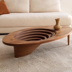 May include: An oval coffee table crafted from dark brown wood, featuring a recessed center with concentric, layered circular cutouts that create a deep, tiered effect. The table stands on a solid, curved base and supports a small, woven vase.