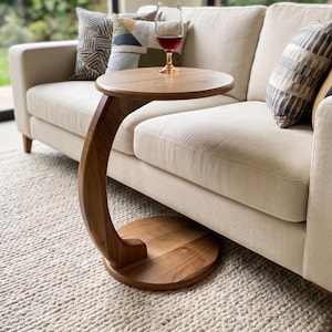 May include: A C-shaped side table crafted from smooth brown wood, featuring a circular tabletop and a matching circular base connected by a curved support column. A clear glass filled with red liquid sits on the tabletop surface.