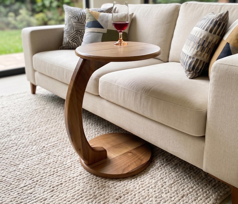 May include: A C-shaped side table crafted from smooth brown wood, featuring a circular tabletop and a matching circular base connected by a curved support column. A clear glass filled with red liquid sits on the tabletop surface.