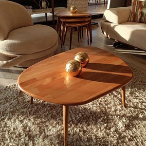 May include: A light brown wooden coffee table with a smooth, rounded triangular top and three tapered legs. Two polished gold spherical decorative objects with intricate swirling patterns are placed on the center of the table surface.