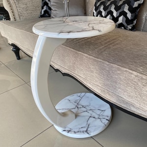 Puede incluir: Una mesa auxiliar en forma de C con un tablero y una base circulares de patrón de mármol blanco con vetas negras. La mesa tiene una columna de soporte curva de color blanco brillante que conecta el tablero y la base, creando una estructura voladiza.