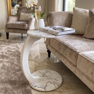 May include: A white C-shaped side table with a faux marble top and base featuring grey veining. The table supports a glass of champagne, a white vase with a white orchid, and a book titled Chanel with a black and white cover photograph.