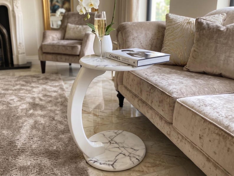 May include: A white C-shaped side table with a faux marble top and base featuring grey veining. The table supports a glass of champagne, a white vase with a white orchid, and a book titled Chanel with a black and white cover photograph.