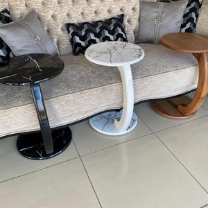 May include: Three C-shaped side tables arranged in a row. One table features a black marble top and base with a black curved support. The center table has a white marble top and base with a white curved support. The third table is made of wood.