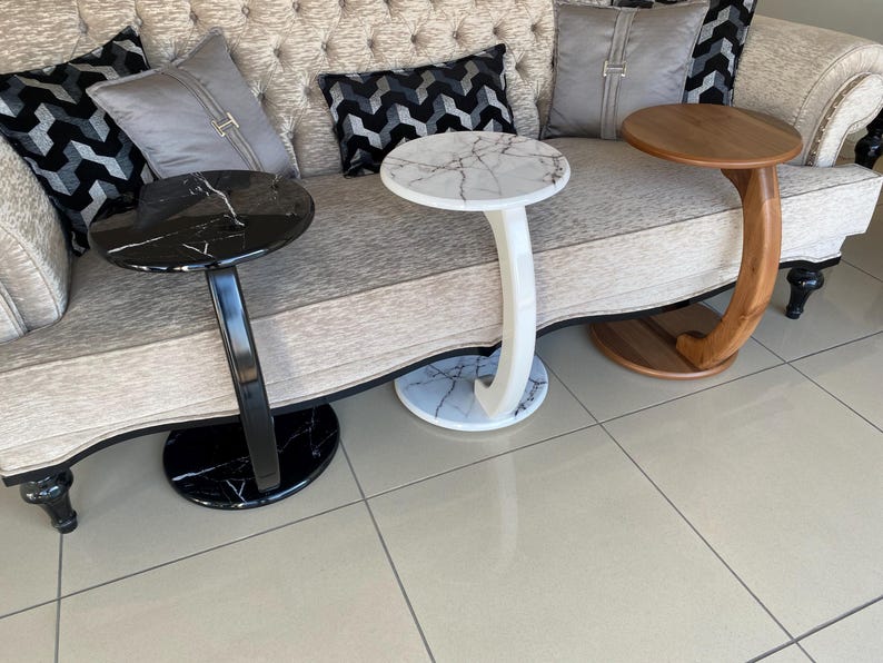 May include: Three C-shaped side tables arranged in a row. One table features a black marble top and base with a black curved support. The center table has a white marble top and base with a white curved support. The third table is made of wood.