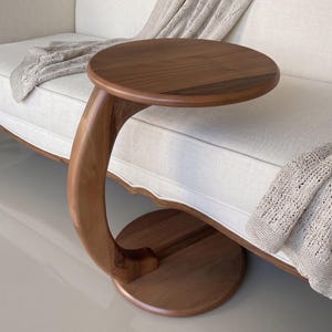 Puede incluir: Una mesa auxiliar en forma de C hecha de madera marrón lisa, con un tablero circular y una base circular a juego conectados por una columna de soporte curva que permite deslizar la mesa sobre el borde de un sofá o una silla.
