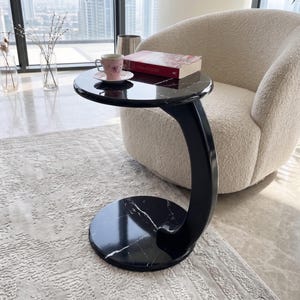 May include: A C-shaped side table with a black marble top and base featuring white veining. The table has a curved black support column connecting the circular top and base. A pink floral teacup and a red book are placed on the table surface.