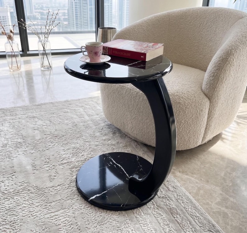 May include: A C-shaped side table with a black marble top and base featuring white veining. The table has a curved black support column connecting the circular top and base. A pink floral teacup and a red book are placed on the table surface.