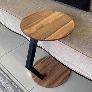 May include: A C-shaped side table featuring a circular wooden top and a matching circular wooden base connected by a single, angled black metal support frame. The wood displays a natural grain pattern in shades of medium brown and dark brown.