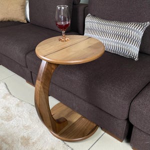 May include: A C-shaped side table crafted from natural brown wood with a circular tabletop and a matching circular base connected by a curved support column. A clear glass filled with red liquid sits on the smooth wooden surface of the table.