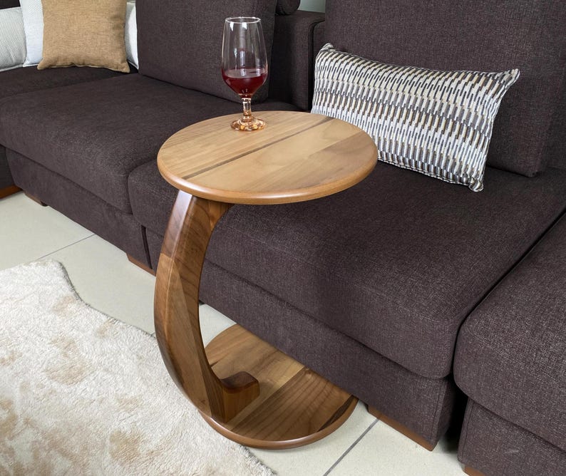May include: A C-shaped side table crafted from natural brown wood with a circular tabletop and a matching circular base connected by a curved support column. A clear glass filled with red liquid sits on the smooth wooden surface of the table.
