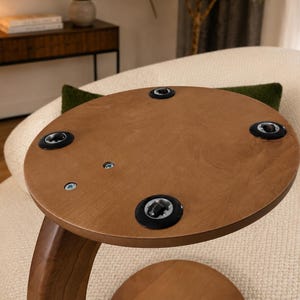 May include: A round, light brown wooden tabletop viewed from the underside, featuring four black circular plastic inserts positioned near the edge and two exposed metal screws embedded in the wood surface near the center of the circular panel.