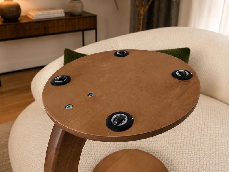May include: A round, light brown wooden tabletop viewed from the underside, featuring four black circular plastic inserts positioned near the edge and two exposed metal screws embedded in the wood surface near the center of the circular panel.